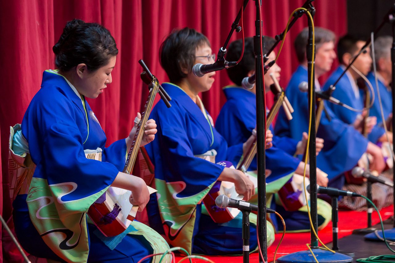 People wearing colorful kimonos sitting on a stage playing instruments; curtain; microphones