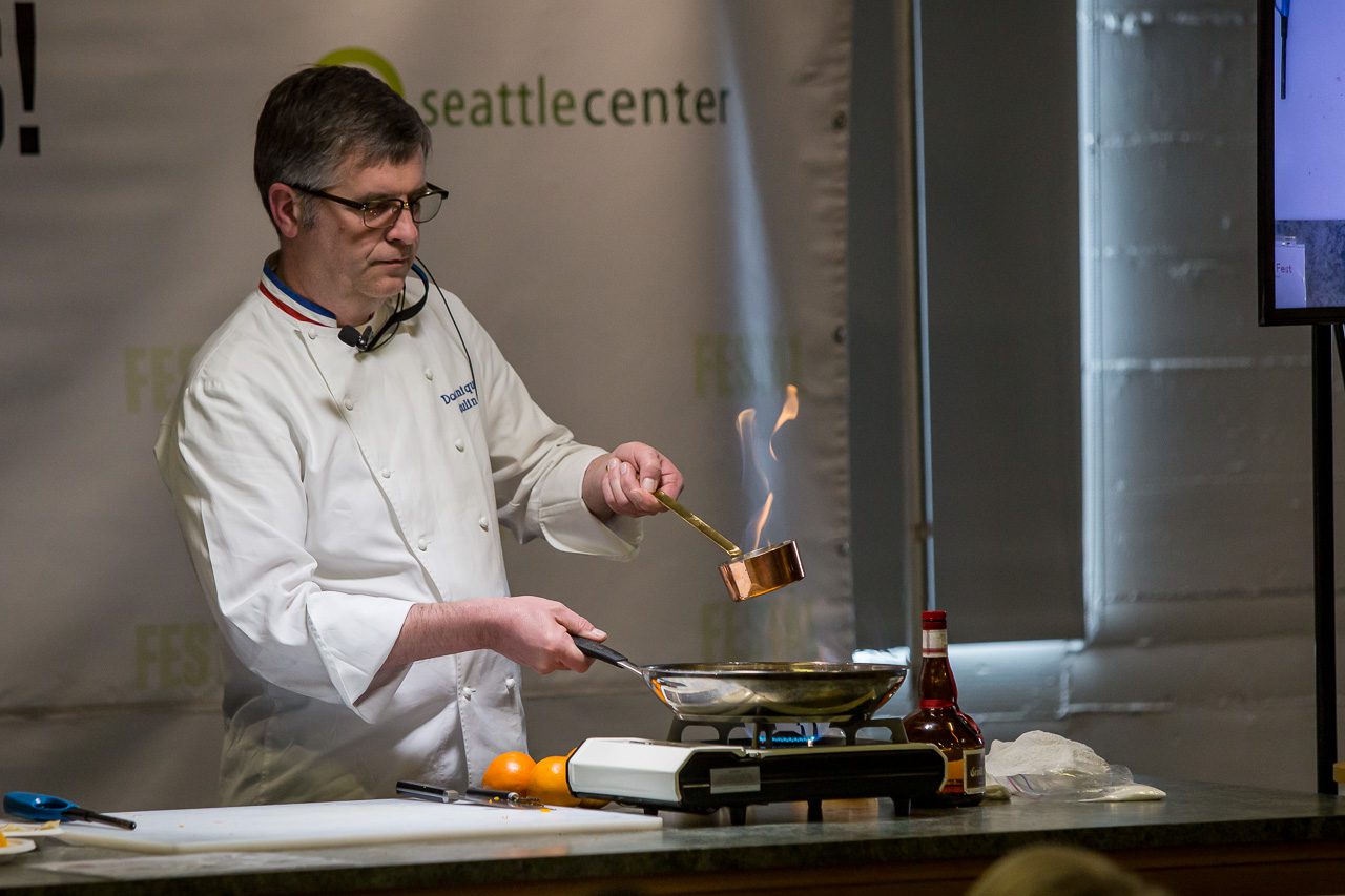 A man (who is a chef/cooking expert) who wears glasses, is demonstrating French cooking techniques. He is holding a pan handle in his right hand and in his left hand and in left hand he is holding a ladle that has a flame on top. 
Pictured in the photo: pan, electric burner, cutting board, Grand Marnier Liqueur, oranges, lighter, knife, tabletop, banner that says "Seattle Center"