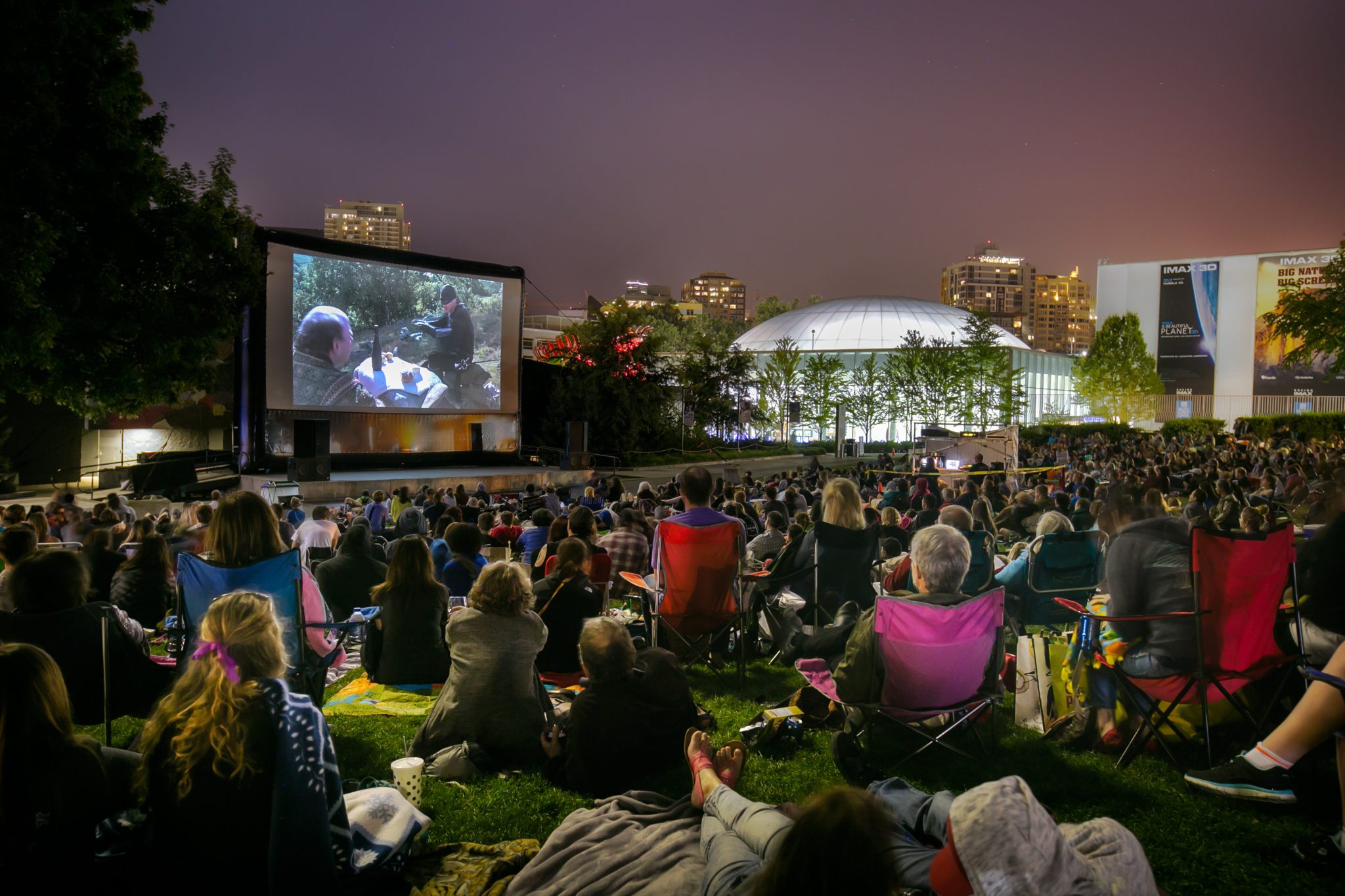 Outside movie screen, people sitting on blankets, chairs, grass, trees, buildings, night time