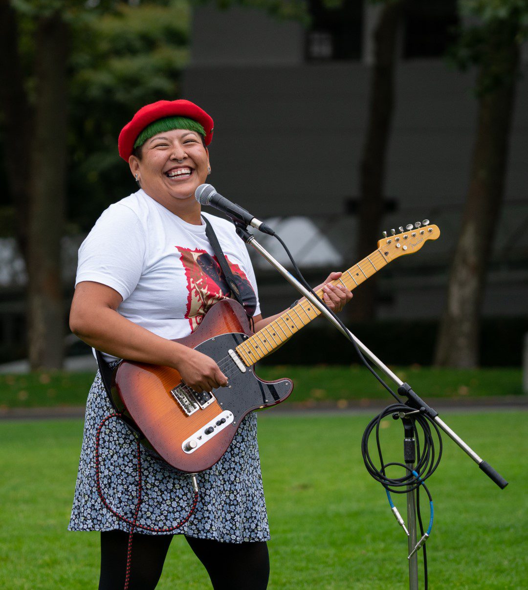 A woman playing an electric guitar, microphone stand; grass; trees; building in the background; daytime
