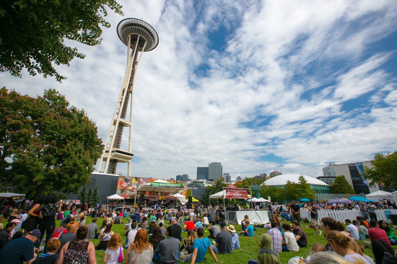 A crowd sitting and standing in a grassy area. Food vendors, buildings, trees, clouds, day time.