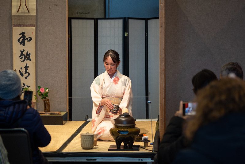 Tea ceremony, people gathered around a woman seated on a grass mat, holding a cup. 