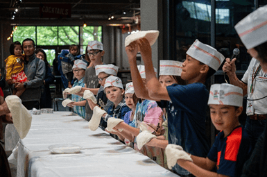 A group of kids at a table learning to spin pizza dough